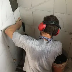 This image shows Brother Grimes installing tile in a bathroom renovation. He's wearing red ear protection headphones while carefully placing white wall tiles. A red bucket sits on the floor beside him, likely containing tiling materials. His light-colored shirt shows signs of the hands-on work involved in bathroom remodeling.